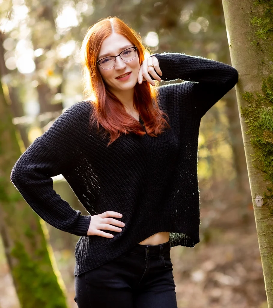 FinaFoxy lehnt mit schwarzem Pullover an einem Baum im Wald