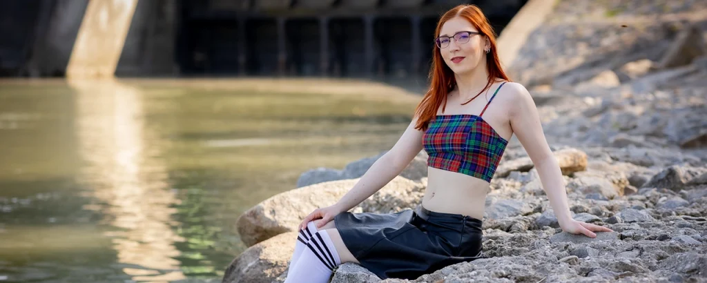FinaFoxy sitzt am Ufer mit einem bauchfreien Top, einem kurzen Rock und Kniestrümpfen